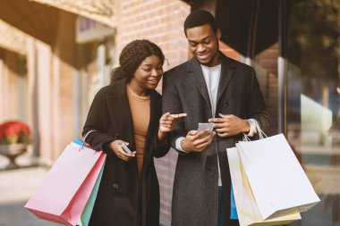 Satın alma için mobil uygulama, online sipariş ve nakit para. Bir sürü renkli alışveriş torbası taşıyan gülümseyen Afro-Amerikan çifti akıllı telefonlara bakar ve açık havada, dükkanın yanında kağıt poşetler tutarlar.