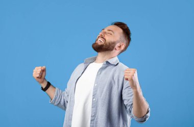 Muzaffer genç adam, mavi stüdyo arka planında kapalı gözlerle EVET işareti yapıyor, panorama. Sakallı milenyum adamı zaferini kutlarken büyük bir başarıyla mutlu hissediyor, şansına inanamıyor