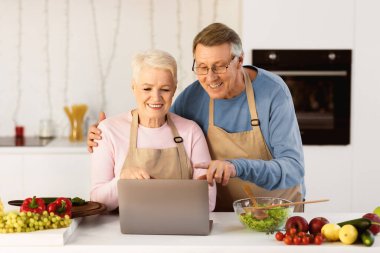 Son sınıf aşçıları internete girip online yemek tarifi ararken ve evde modern mutfakta birlikte sağlıklı bir akşam yemeği hazırlarken. Gıda Hazırlama ve Bilgisayar Kullanımından Hoşlanan Yaşlı Eşler