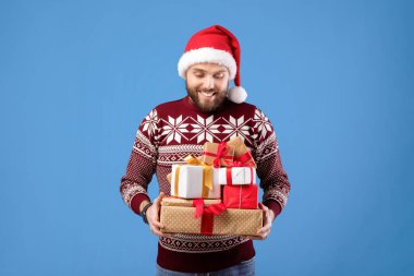 Mavi stüdyo arka planında Noel Baba şapkası ve Noel Baba şapkalı mutlu beyaz adamın portresi. Noel 'i ya da yeni yılı kutlayan hediye yığınıyla yakışıklı genç adam.