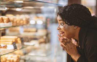 Harika kekler ve tatlıya düşkünlük konsepti. Şaşırmış, mutlu Afro-Amerikan milenyum bayanı ağzı açık bir şekilde modern şekerci dükkanında şekerleme dükkanının vitrinine bakıyor, kapanıyor, boşluğu kopyalıyor.