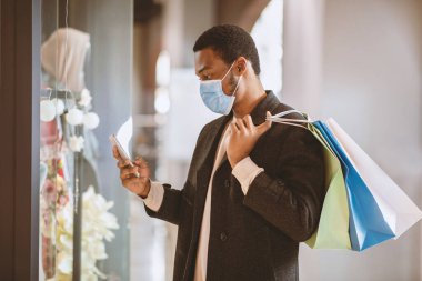 Yeni normal, COVID-19 salgını sırasında Kara Cuma 'da alışveriş ve satış. Paltolu ve koruyucu maskeli, ciddi bir Afro-Amerikan erkeği renkli çantalar tutuyor ve dükkan penceresinin yanındaki akıllı telefondan bakıyor.