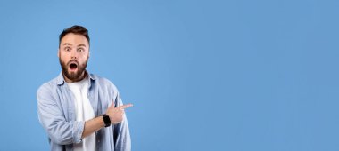 Şok olmuş genç adam, ağzı açık duruyor, inanamadığı boşluğu işaret ediyor, mavi stüdyo arka planı, afiş tasarımı. Şaşkına dönmüş sakallı adam büyük bir satış ya da terfi teklif ediyor.