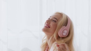 Genç sarışın bir kadın sabah evde müzik dinlerken neşeyle gülümsüyor. Güneş ışığı perdelerin arasından süzülüp sıcak ve davetkar bir atmosfer yaratır..