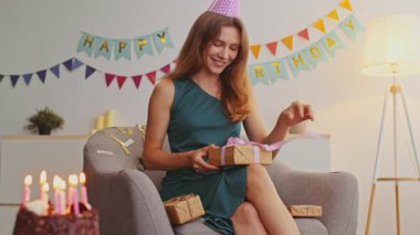 Kızıl saçlı genç bir kadın evde doğum günü kutlaması yapıyor. Bir sandalyede oturuyor, hediyeyi açarken gülümsüyor, etrafı şenlikli süslemelerle ve mumlu pastayla çevrili..