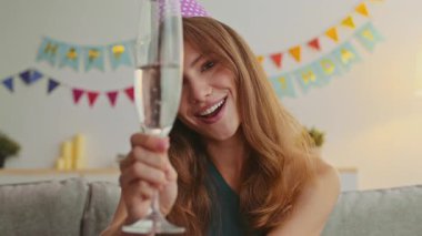 Kızıl saçlı genç bir kadın evinde bir bardak köpüklü içecekle mutlu bir şekilde doğum gününü kutluyor. Renkli dekorasyonlar, o çevrimiçi olunca şenlikli bir atmosfer yaratıyor..