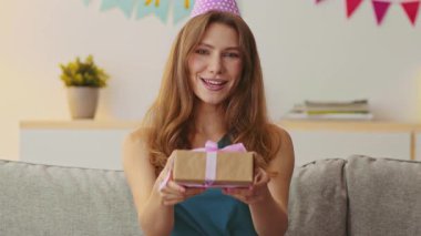 Kızıl saçlı beyaz bir kadın evde doğum gününü kutluyor. Bir parti şapkası takıyor ve elinde güzel paketlenmiş bir hediye tutuyor, süslerle çevrili özel gününün tadını çıkarmaya hazır..