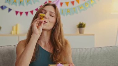 Genç, kızıl saçlı bir kadın evde doğum gününü kutluyor. Elinde bir hediyeyle ışıl ışıl gülümsüyor. Renkli dekorasyonlar arkaplanı dolduruyor, şenlikli atmosfere ekliyor.