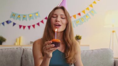 Kızıl saçlı genç bir kadın, elinde mumla elinde kek tutarak mutlu bir şekilde doğum gününü kutluyor. Renkli dekorasyonlar ve sıcak bir atmosfer şenlik havasını artırır..