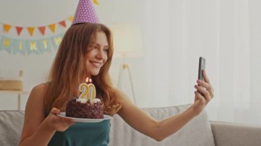 Evde kutlama yaparken parti şapkası takan neşeli genç bir kadın doğum günü pastasını gösteriyor. Özel anını paylaşmak için arkadaşlarıyla video görüşmesi yapmaktan hoşlanıyor..