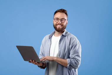 Gözlüklü neşeli bir milenyum adamı online iş için dizüstü bilgisayar kullanıyor ya da mavi stüdyo arka planında iletişim kuruyor. Yakışıklı genç adam video izliyor, Webinar 'a katılıyor.