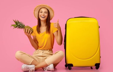Yazlık elbiseli, ananaslı mutlu bir kız başparmağını kaldırıyor, pembe stüdyo arka planında bavulun başında oturuyor.