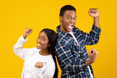 Zaferin duygularını ifade etmek ve zaferi birlikte kutlamak. Bin yıllık Afro-Amerikan erkeği ve bayanı sırt sırta verin ve yumruklarınızı kaldırın, sarı arka planda izole edilmiş, stüdyo çekimi, kopyalama alanı