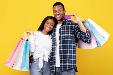 Alışveriş, boş zaman ve mevsimlik satış. Neşeli, genç, Afro-Amerikan çift birbirlerine aşık. Renkli çantalar, sarılma ve gösteriş kartları, sarı arka planda izole edilmiş, stüdyo çekimi, fotokopi alanı.