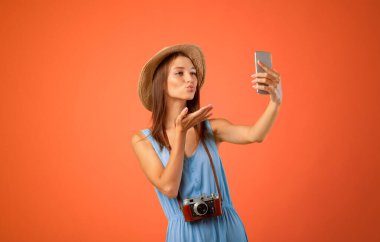 Mutlu genç bayan gezgin cep telefonuyla selfie çekiyor, sarı stüdyo geçmişi var. Yazlık şapkalı neşeli kadın sosyal medya için fotoğraf çekiyor, akıllı telefon kullanıyor, uçan öpücük yolluyor.