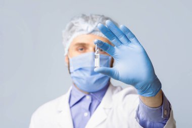 Tıbbi maske takan genç bir erkek doktor ve koruyucu eldivenler gri stüdyo arka planında aşı ya da ilaç bulunduruyor, seçici odaklanma. Viral hastalığın yenilikçi tedavisini gösteren doktor