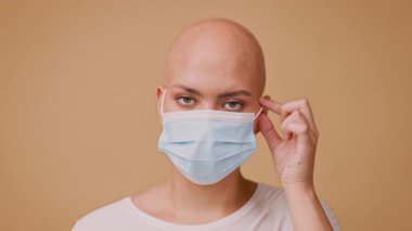 Kel kafalı genç bir kadın, arka planda mavi bir maske takıyor. Yüzü kameraya dönükken, kendine güveni tam, benzersiz görünüşünü ve tarzını sergiliyor..