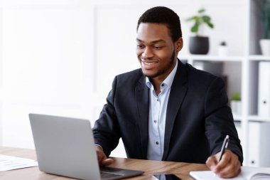 Neşeli genç siyah iş adamı dizüstü bilgisayarda webinar dinliyor ve notlar alıyor, ofis içi, fotokopi alanı. Gülümseyen Afrikalı Amerikalı yönetici dizüstü bilgisayar kullanıyor ve not defterine bir şeyler yazıyor.