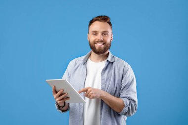 Modern teknolojiler ve iletişim. Mavi stüdyo geçmişi üzerinde tablet bilgisayar kullanan mutlu sakallı adamın portresi. Dokunmatik bloknotlarda çalışan ya da okuyan genç adam.