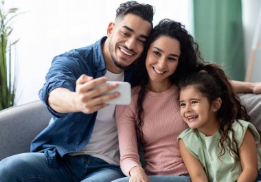 Aile selfie 'si. Arap bir adam, evinde karısı ve küçük kızıyla akıllı bir telefonla fotoğraf çekiyor. Oturma odasında koltukta otururken mutlu bir orta doğu aile pozu veriyor.