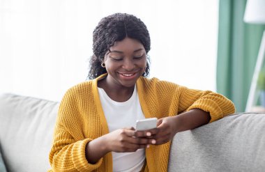Akıllı telefonlu güzel Afro-Amerikan bayan hafta sonu evde eğleniyor, yakın plan portresi. Gülümseyen siyah bir kadın arkadaşlarıyla ya da sevgilileriyle sohbet ediyor, cep telefonu kullanıyor, oturma odasını kullanıyor.