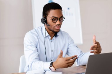 Afrika kökenli Amerikalı bir adam bilgisayar, ofis içi ve fotokopi alanı üzerinden iş ortaklarıyla görüntülü görüşme yapıyor. Yakışıklı siyah menajer ya da iş koçu iş yerinden yayın yapıyor.
