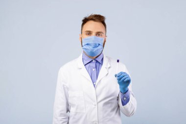 Koruyucu maskeli Millennail erkek doktor ve steril çubuk tutan eldivenler Coronavirus PCR testi için gri stüdyo arka planında. Geniz ve oral covid tanı prosedürü için tüplü genç doktor.