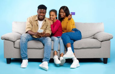 Online Shopping Concept. Happy Young Black Parents With Daughter Using Cell Phone And Debit Credit Card Buying Goods In Internet Web Store, Three People Sitting On Couch Isolated On Blue Studio Wall