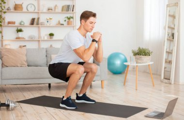 Ev antrenmanı için online koç. Spor giyimli yakışıklı, spor ayakkabılı, spor ayakkabılı, çömelmiş ve oturma odasındaki dizüstü bilgisayara bakan, boş bir yer.
