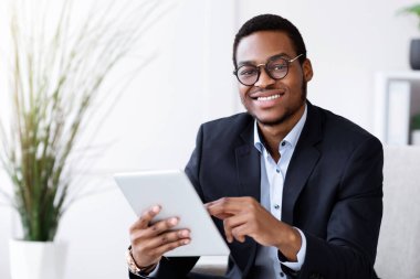 Gülümseyen siyah takım elbiseli genç adam dijital tablet tutuyor, modern teknolojileri iş dünyasında, ofis içinde, kopyalama alanında kullanıyor. Neşeli Afro-Amerikan iş adamı iş için uygulama kullanıyor.