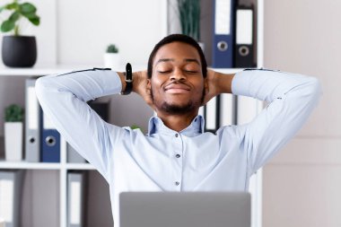 Tatmin olmuş Afro-Amerikan genç işadamı, modern ofisteki dizüstü bilgisayarının önünde rahat bir şekilde, başarılı bir iş anlaşması ya da internet konferansından sonra dinleniyor.