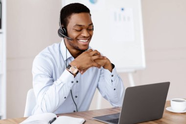 Mutlu siyahi adam çevrimiçi eğitim alıyor, kulaklık ve laptop kullanıyor, bilgisayar ekranına bakıyor ve gülümsüyor, ofis içi, kopyalama alanı. Neşeli Afrikalı adam müşterileriyle online görüşme yapıyor.