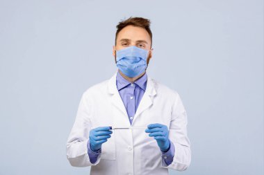 Tıbbi maske ve lastik eldiven takan genç bir doktor gri stüdyo arka planında koronavirüs PCR test tüpü tutuyor. Geniz ve oral covid-19 teşhisi için GP şişesiyle portresi