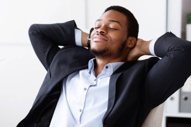 Siyah takım elbiseli, rahat Afro-Amerikan genç iş adamının, çalışma günü ya da başarılı bir iş toplantısından sonra sandalyeye yaslanıp dinlenmesi.