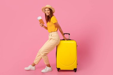 Yazlık giysiler içinde gülümseyen, pembe arka plana yaslanmış, elinde biletlerle pasaport tutan genç bir kadın.