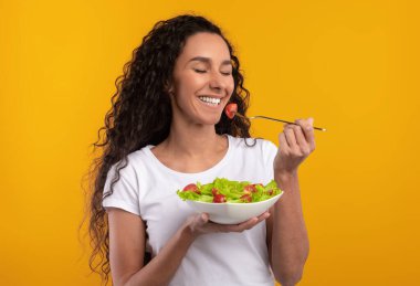 Sağlıklı beslenme kavramı. Lezzetli sebze salatası ve gülümseyen turuncu duvarlı çatal ve kase tutan neşeli, heyecanlı bayan. Mutlu kadın portresi dengeli yemek yiyor, kapalı gözlerle poz veriyor.