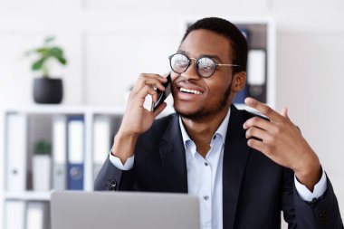 Başarılı iş konuşmaları. İyi ki Afro-Amerikan genç işadamı cep telefonuyla konuşuyor ve modern ofiste el kol hareketi yapıyor, boş alana bakıyor, iş yerinde dizüstü bilgisayarının önünde oturuyor.