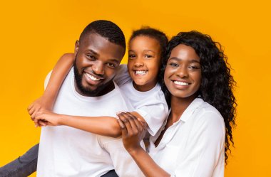 Üç kişilik pozitif siyah bir ailenin portresi, anne, baba ve küçük kızları sarı arkaplanda kameraya bakıyor, boşluğu kopyalıyorlar.