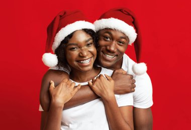 Noel Baba şapkalı genç siyahi aşıklar kırmızı stüdyo arka planında eğleniyor, yakın plan. Neşeli Afrikalı adam mutlu güzel karısına arkadan sarılıyor, 2021 yılını ya da Noel 'i birlikte kutluyor.