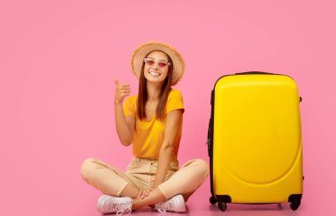 Yazlık şapkalı ve güneş gözlüklü neşeli genç bir kadın bavullarıyla yerde oturuyor ve fotokopi odasını işaret ediyor. Pembe stüdyo arka planı. Yaz kıyafetleri içindeki mutlu bayan heyecan verici seyahat anlaşmaları gösteriyor.