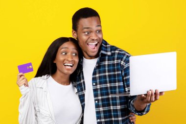 Harika alışveriş, büyük indirim, büyük indirim. Neşeli kahkahalar atan binlerce yıllık Afro-Amerikan erkeği, dizüstü bilgisayara bakıyor, kadın kredi kartı tutuyor, sarı arka planda izole edilmiş, stüdyo çekimi, boş yer.