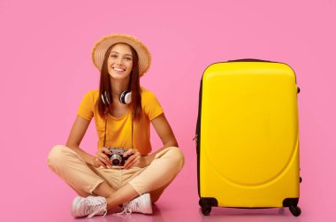 Pembe stüdyo arka planında bavulun başında oturan neşeli genç kadın. Yazlık şapkası, kulaklığı ve kamerasıyla gezmeye hazır gülümseyen bayan. Aktif dinlenme, tatil, turizm, seyahat kavramı