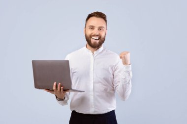 Resmi olarak heyecanlı bir ofis çalışanı dizüstü bilgisayar tutuyor ve gri stüdyo geçmişi üzerinde EVET jestleri yapıyor. Çevrimiçi çalışan olumlu genç adam, başarısını ya da iş başarısını kutluyor.