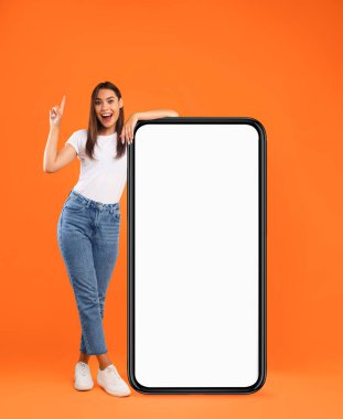 Mobil Uygulama Reklamı, Harika Teklif. Heyecanlı Bayan Parmak Gösterme Uzunluğu Turuncu Stüdyo Duvarı 'nda duran kocaman beyaz akıllı telefon ekranına yaslanıyor. Cep telefonu Görünümü Modifiye Et