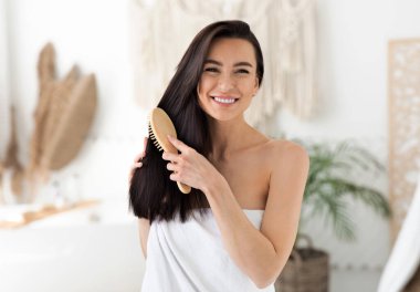 Kadınlar boş vakitlerinde evde saç bakımını, tedaviyi ve spa gününü yaparlar. Havluya sarılmış, uzun ve sağlıklı saçlarını tarak ile tarayan mutlu, genç, tatlı esmer kadın.