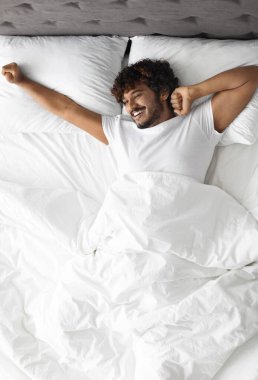 Beyaz tişörtlü mutlu kıvırcık Hintli adam rahat yatakta uzanıp fotokopi odasına bakıyor ve gülümsüyor, yeni günün tadını çıkarıyor, tamamen dinlenmiş hissediyor.