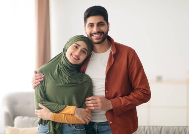 Sevgili Orta Doğulu çift evde poz veriyor. Müslüman koca kameraya gülümseyen tesettürle karısına sarılıyor. Aşk ve Aşk, Mutlu İlişkiler ve Evlilik Konsepti