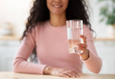 Sağlıklı içecek. Mutfakta masada otururken mineralli suyla gülümseyen kadın, evde su içen esmer kadın, seçici bir odak noktasıyla yakın plan çekilmiş.