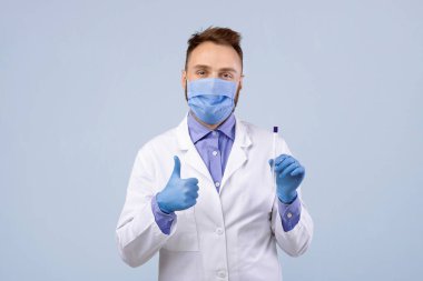 Maskeli ve eldivenli genç doktorun portresi elinde kovid-19 PCR test tüpü ve gri stüdyo arka planında baş parmak hareketlerini gösteriyor. Sağlık çalışanı koronavirüs tanı prosedürünü tavsiye ediyor.