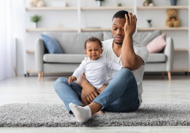 Yorgun, genç bir baba evde yerde ağlayan yeni doğmuş bebekle oturuyor, umutsuz Afrikalı Amerikalı baba bebeğini üzüyor, babalık izni sırasında acı çekiyor.
